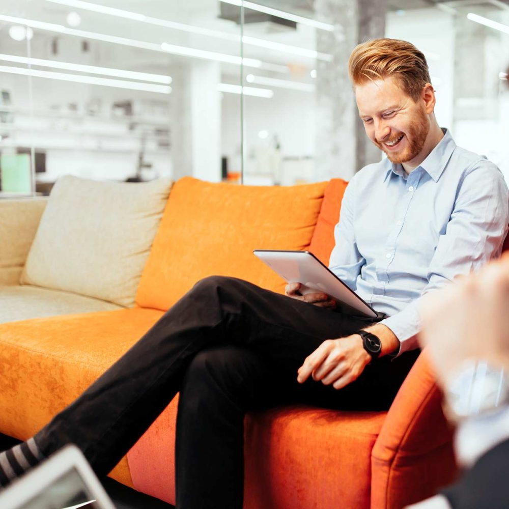 A bearded man dressed in office wear sitting on an orange sofa, looking at an iPad and smiling